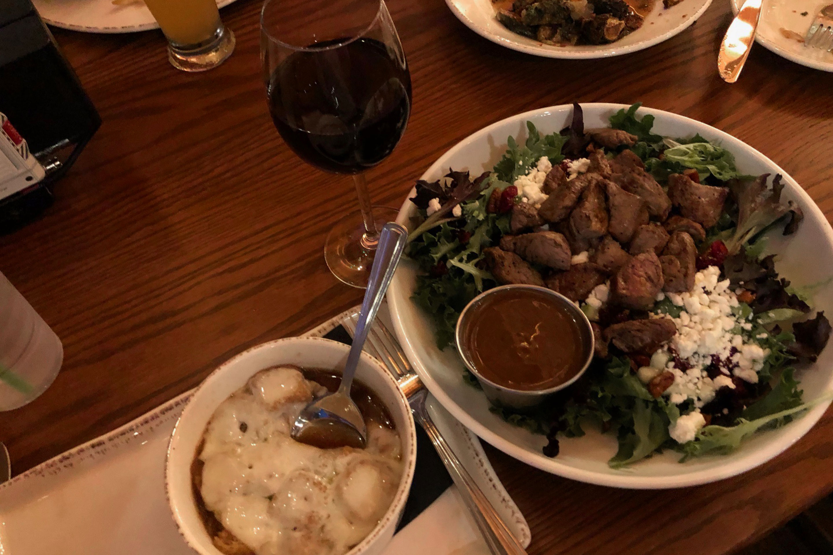 French Onion Soup (left), a glass of red wine (middle) and salad filled with greens, apples, caramelized pecans, cranberries and cheese on a wood table.