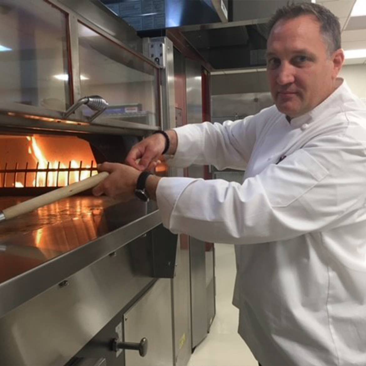 A man wearing a chef jacket stands near an oven.