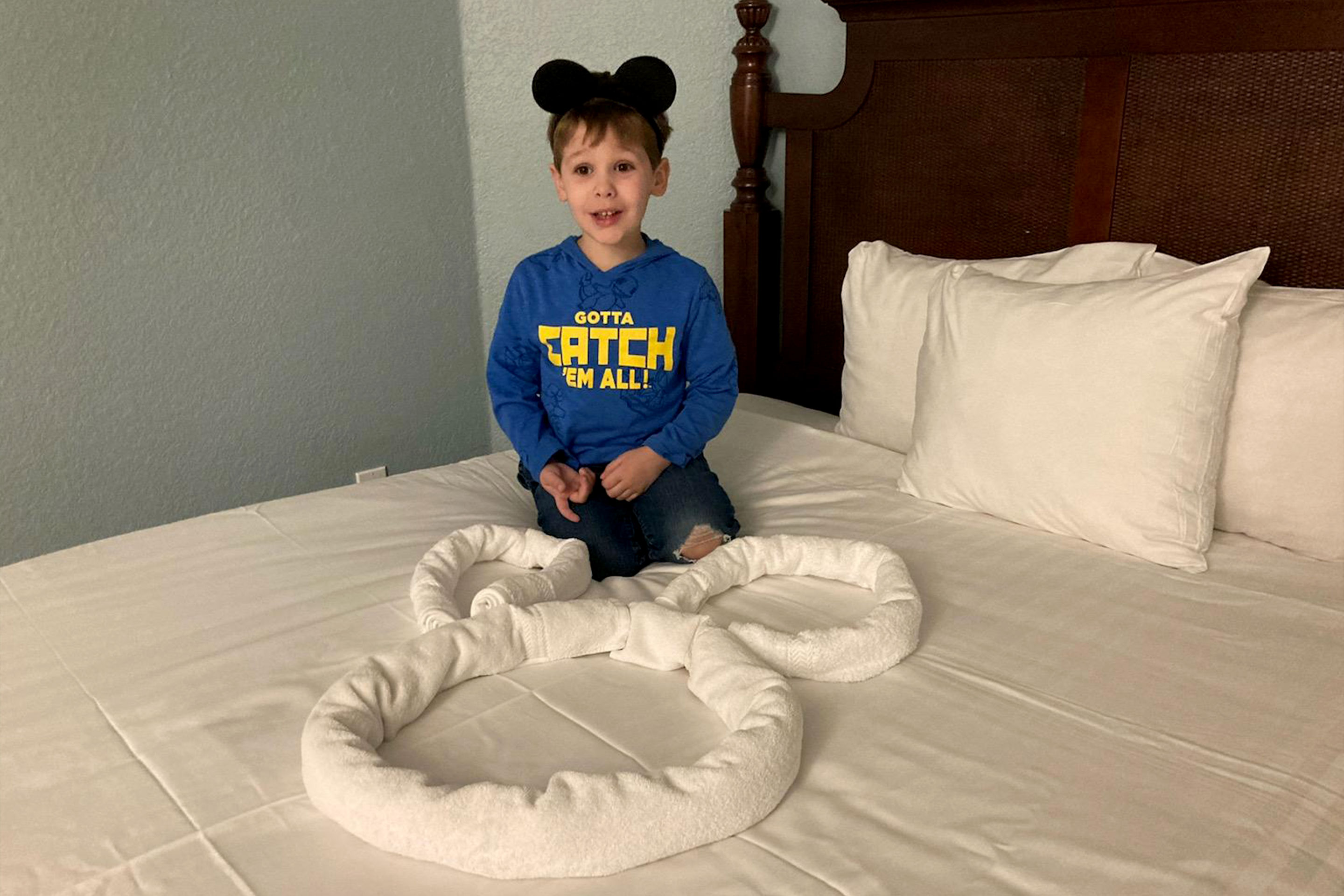 A young boy wearing a blue hoodie with black mouse ears sits on a bed with white linens and three white towels shaped into a Mickey Mouse head.