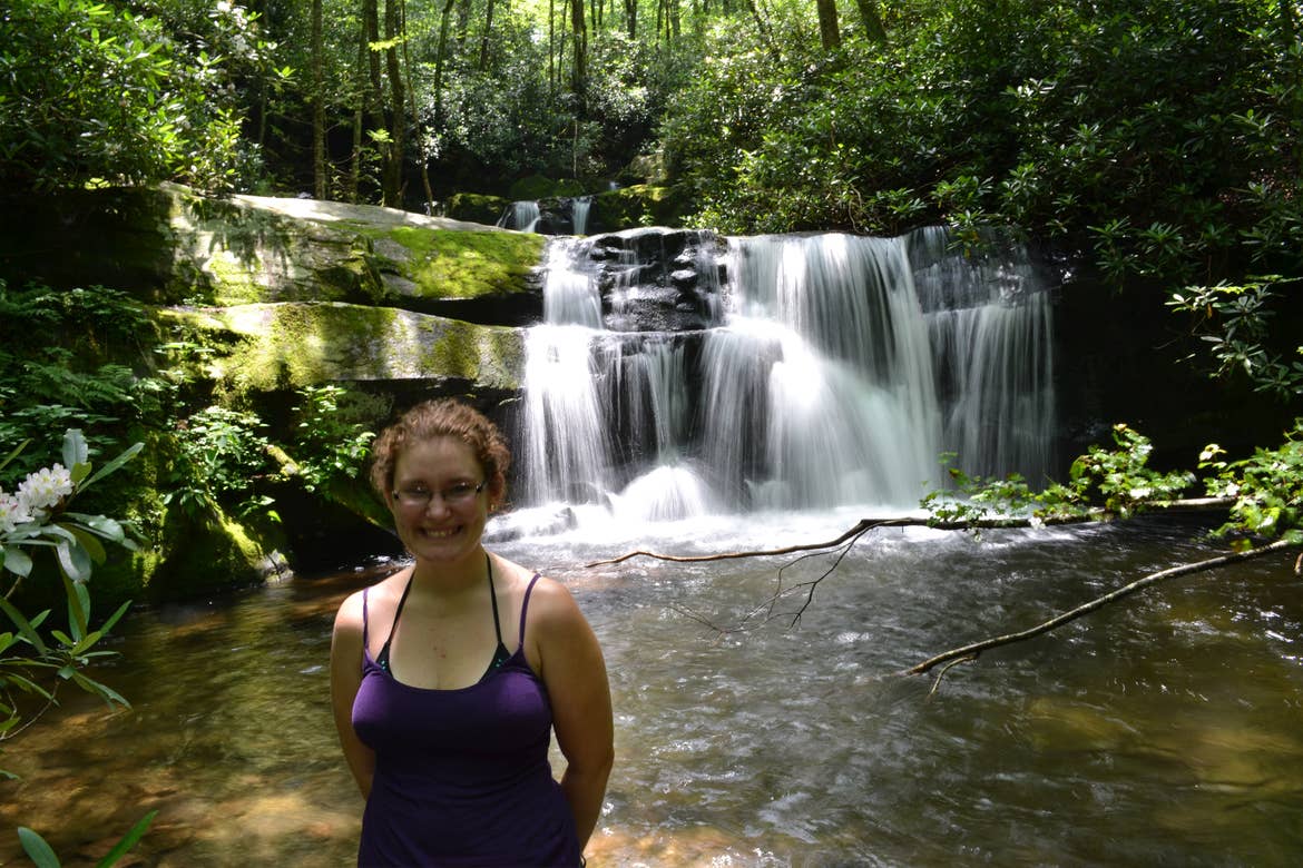 A woman wearing a purple tank top, jean shorts and glasses poses near waterfall cascades over a rock formation at Lynn Camp Prong Cascades in Tennessee.