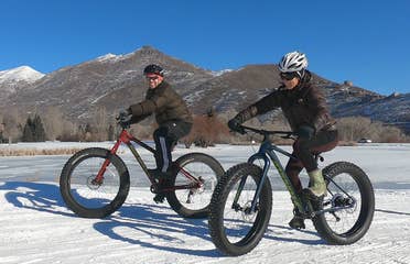 Featured Contributor, Jessica Averett (right), wears a winter jacket, sunglasses, and white bike helmet while riding a Fat Tire bike in the snow next to a man in front of the mountains.