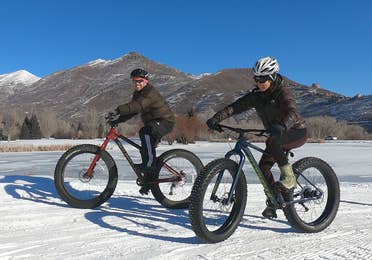 Featured Contributor, Jessica Averett (right), wears a winter jacket, sunglasses, and white bike helmet while riding a Fat Tire bike in the snow next to a man in front of the mountains.