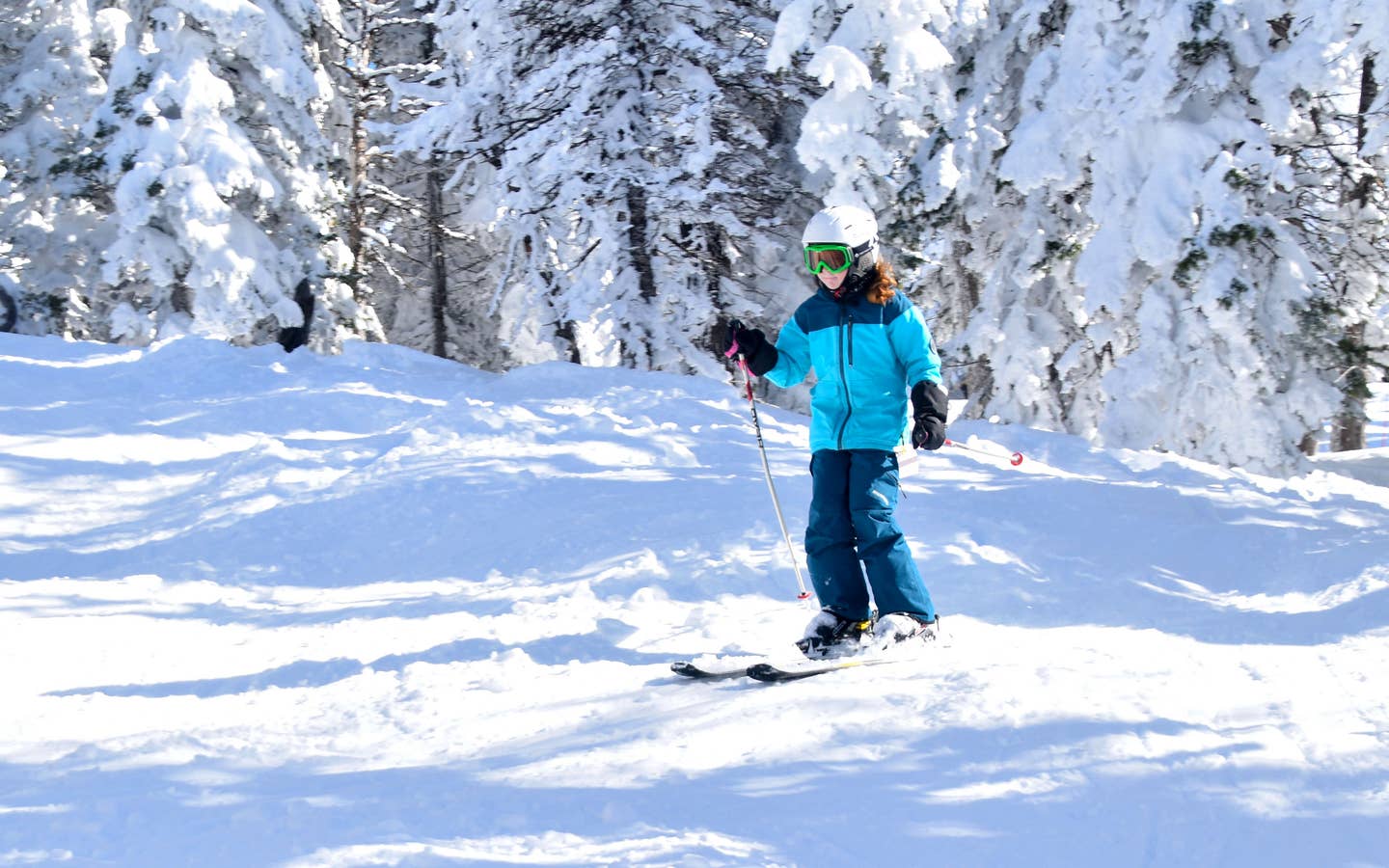 A young girl in blue winter jacket, snow pants and white helmet goes skiing outdoors.