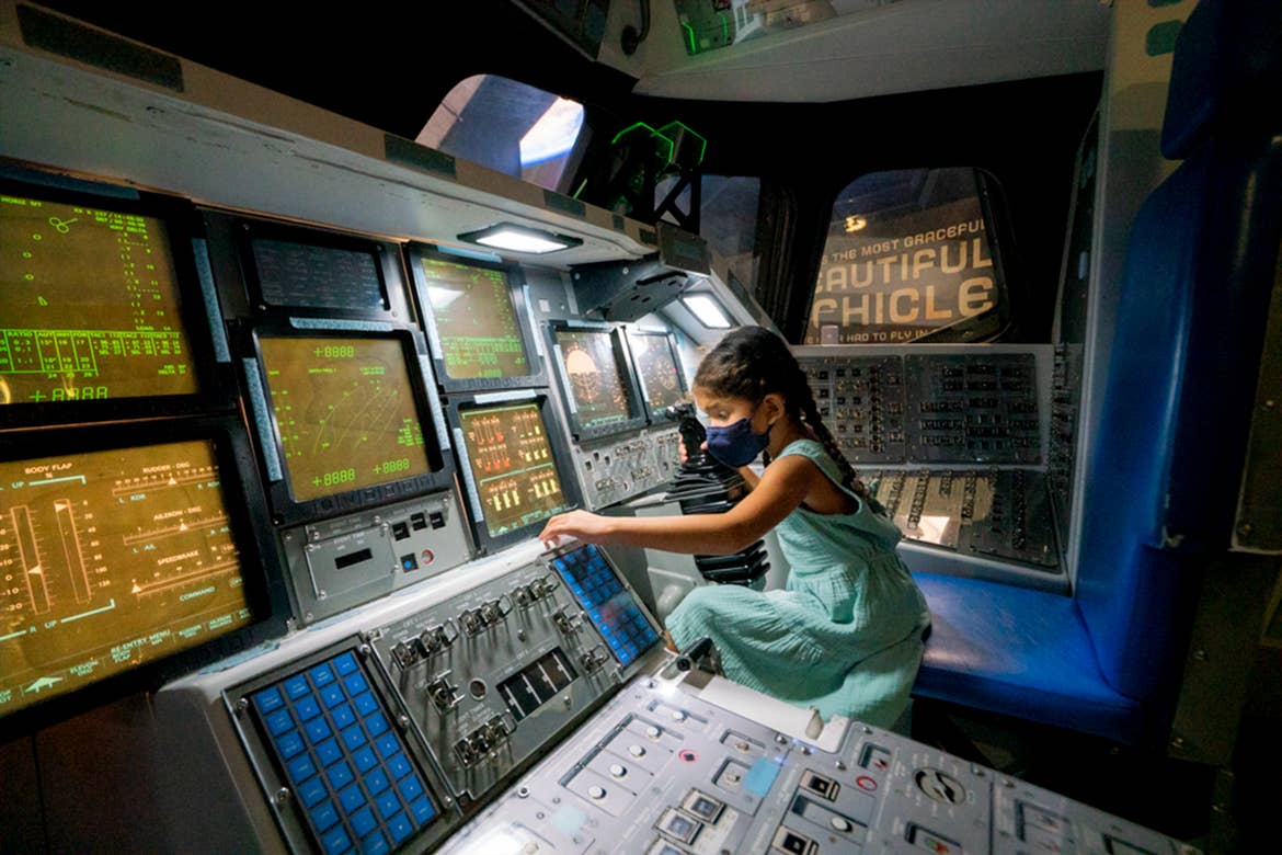A young girl wearing a blue dress and black face mask sits in a rocket ship cockpit playing with controls.