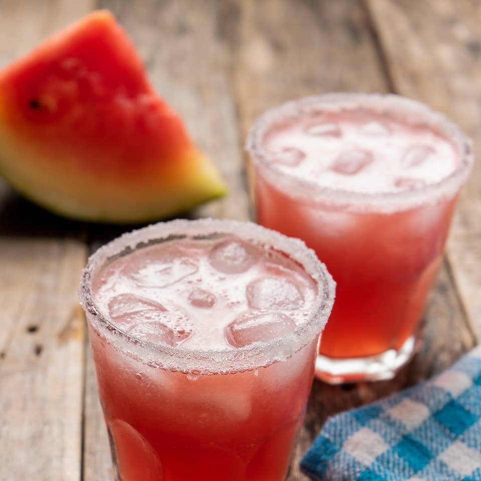 Two glasses of Melon Margaritas sit on a wooden plank table in front of a watermelon wedge.