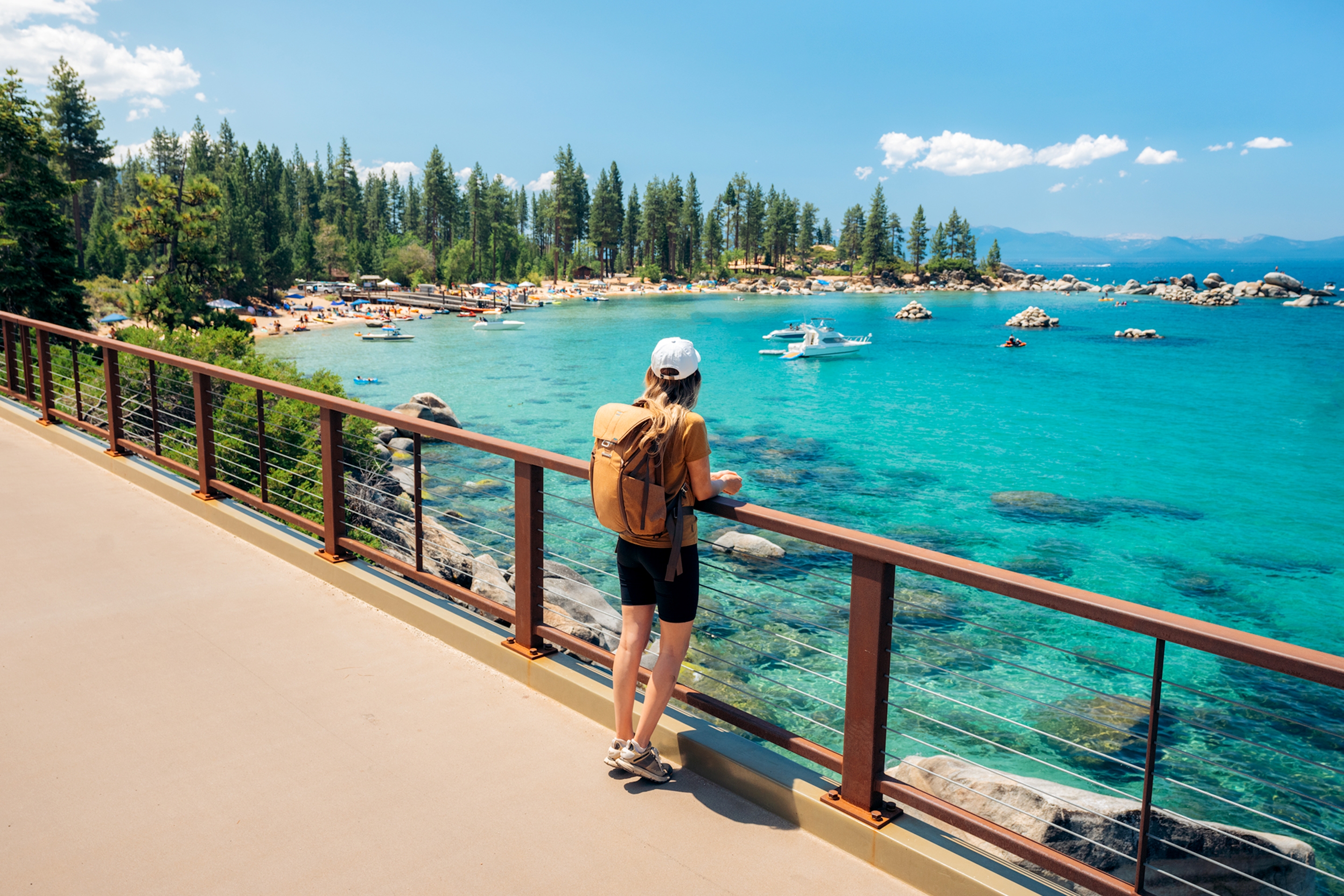 A caucasian woman wearing a mustard shirt, mustard backpack, white baseball cap, black biker shorts and boots leans on a rail near a paved walkway near a lake surrounded by pine trees on a rocky shoreline.