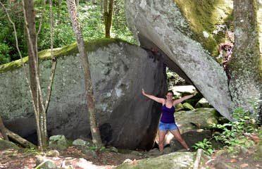 A woman wearing a purple tank top, jean shorts and glasses poses underneath a pair of wedged rocks in Gatlinburg, TN.
