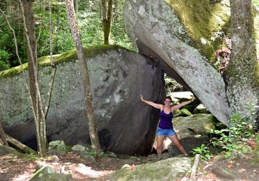 A woman wearing a purple tank top, jean shorts and glasses poses underneath a pair of wedged rocks in Gatlinburg, TN.