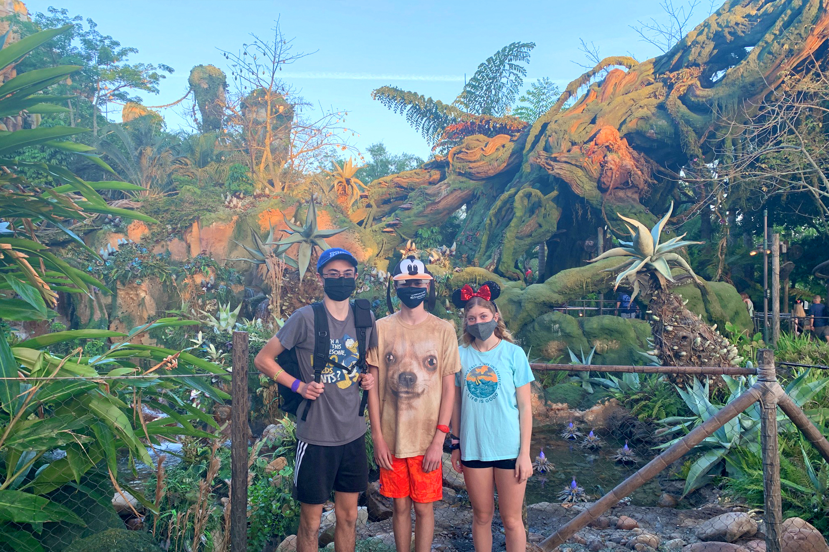 Two young men (left) and a young woman (right) wear Disney hats and safety masks near various greenery in PANDORA - World of Avatar in Disney's Animal Kingdom at Walt Disney World Resort.