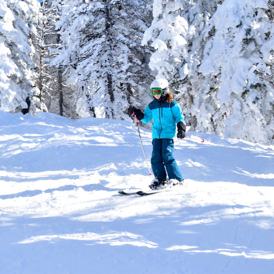 A young girl in blue winter jacket, snow pants and white helmet goes skiing outdoors.