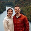 A woman wearing a white pullover, and a man wearing a red puffer jacket stand on a mountain with a river and fall foliage in the background.