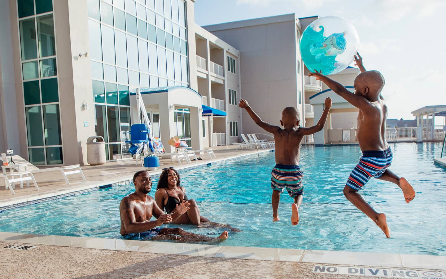 A woman, man and two young boys jump into an infinity pool near a beachfront with a beach ball.