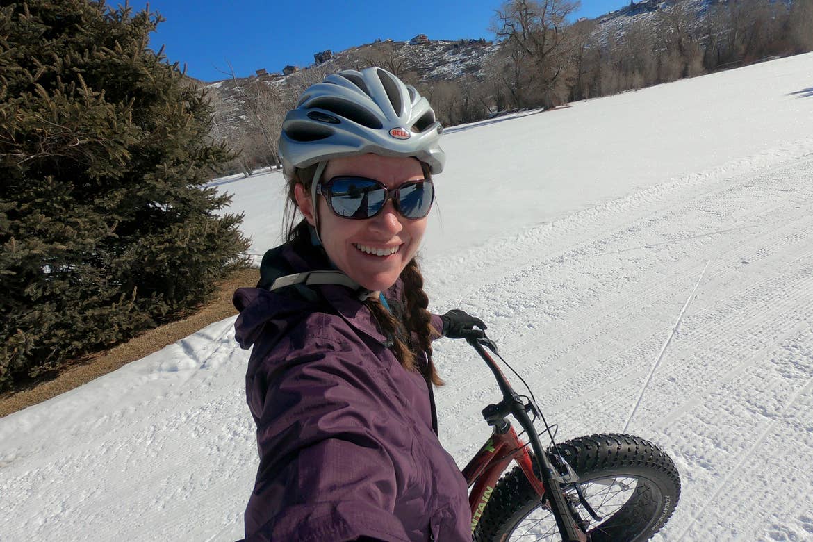 Featured Contributor, Jessica Averett, wears a winter jacket, sunglasses, and white bike helmet while riding a Fat Tire bike in the snow in front of the mountains.
