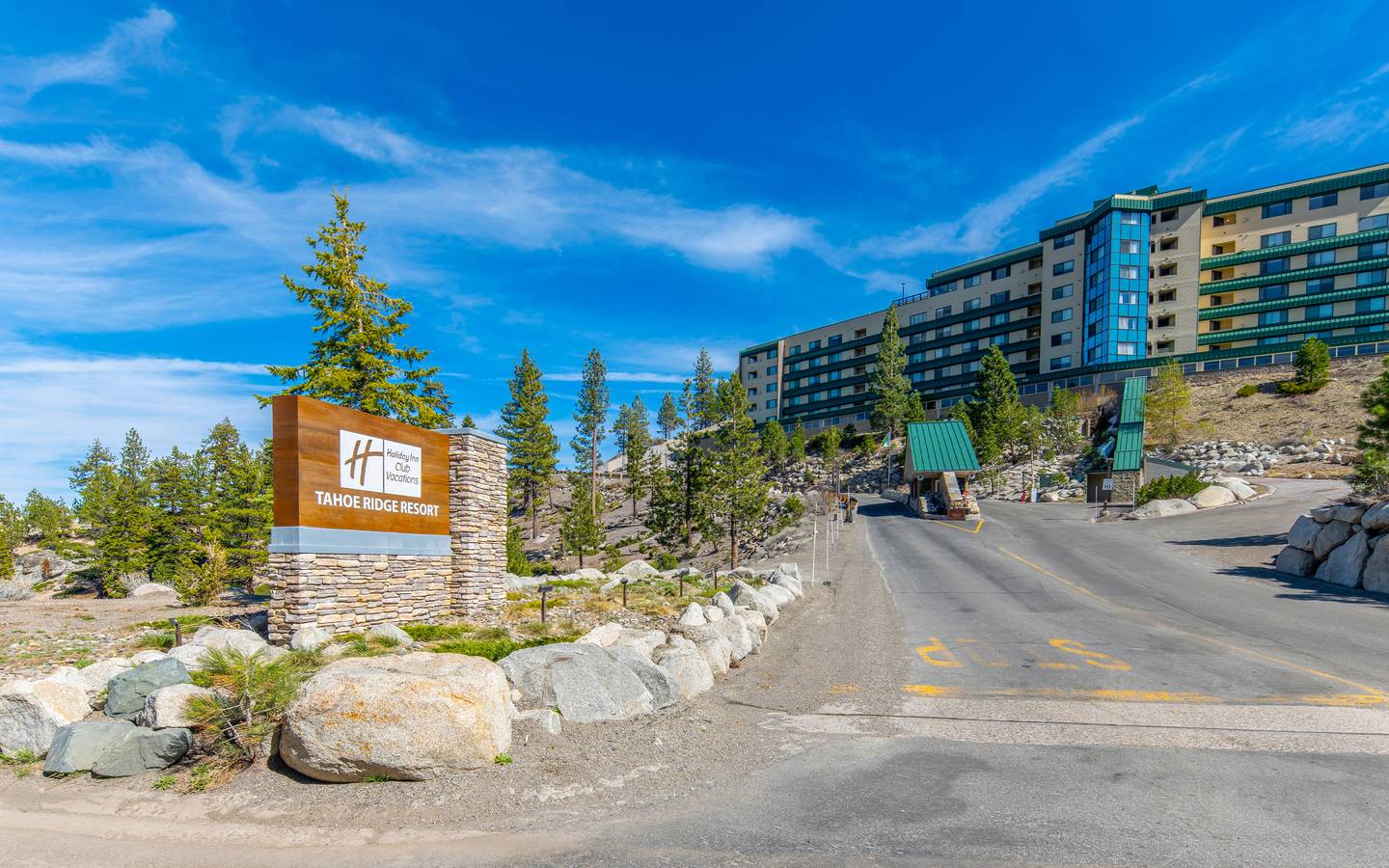 ntrance to Holiday Inn Club Vacations Tahoe Ridge Resort featuring a stone monument sign, uphill driveway with guard gate, and a multi-story resort building set among pine trees under a blue sky.
