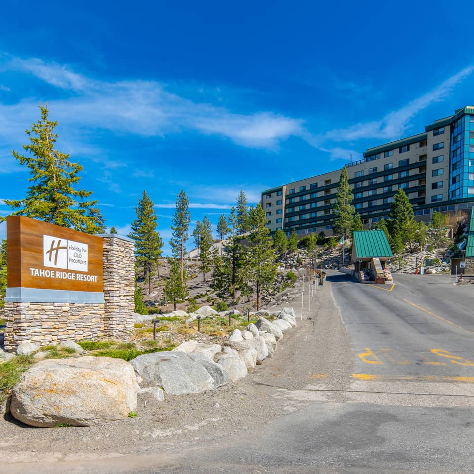 ntrance to Holiday Inn Club Vacations Tahoe Ridge Resort featuring a stone monument sign, uphill driveway with guard gate, and a multi-story resort building set among pine trees under a blue sky.