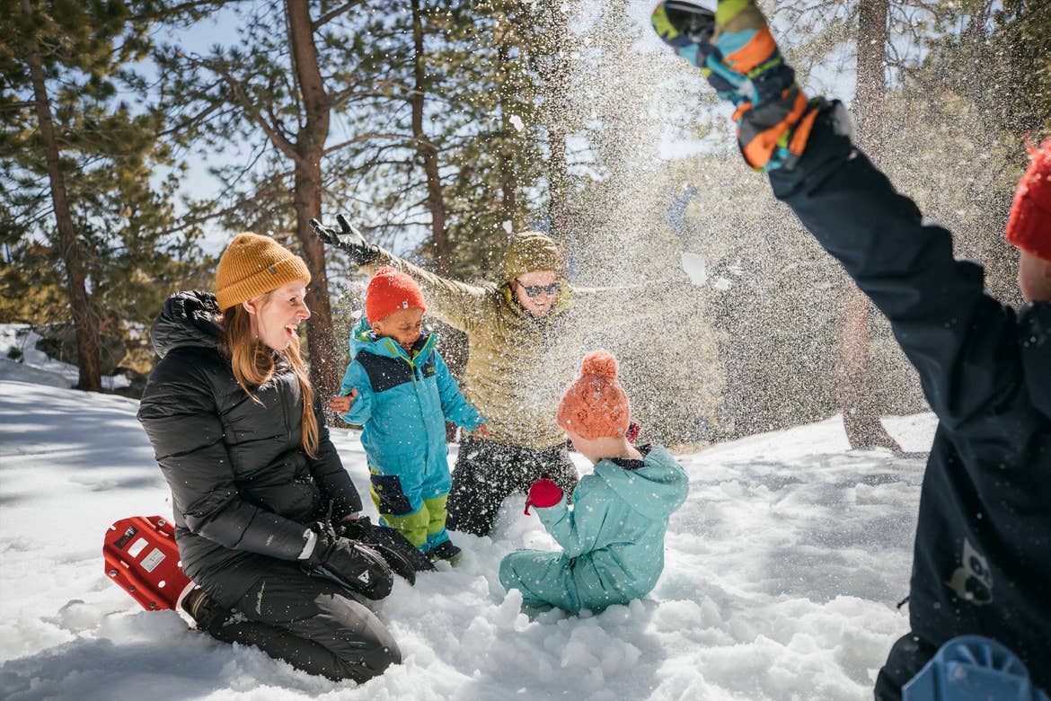 A woman, man and three children wear snow apparel while playing in snow on a mountain.