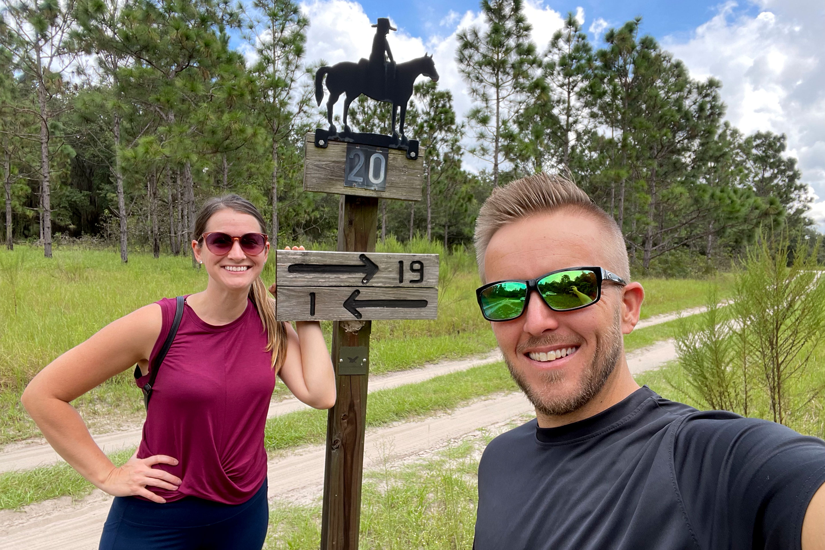 A woman in a red shirt, black pants and sunglasses stands next to a sign near a man with a black shirt and sunglasses outdoors.