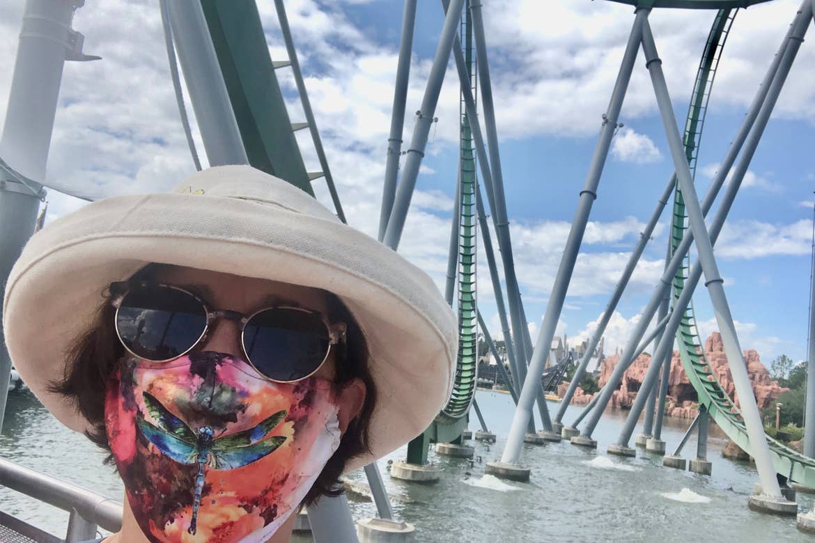Author, Rona, stands in front of the inversions of a rollercoaster and water feature wearing a sunhat and mask.
