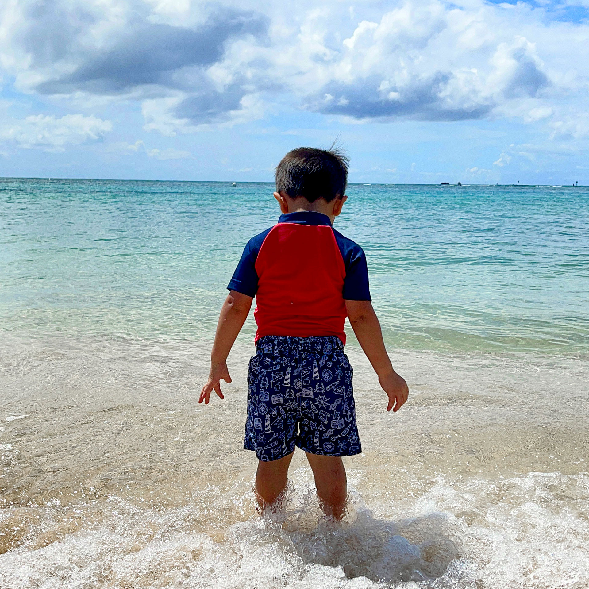 Featured Contributor, Danny Pitaluga's son, Joey, stands in the sand on the beach in swimwear as waves roll in.