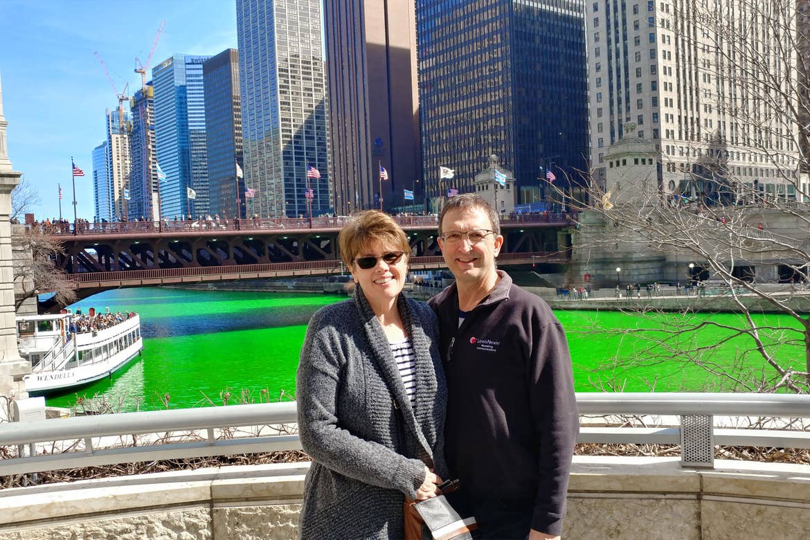 A woman in a grey cardigan and man in a black pullover stand in front of a green-dyed Chicago River and Chicago buildings.