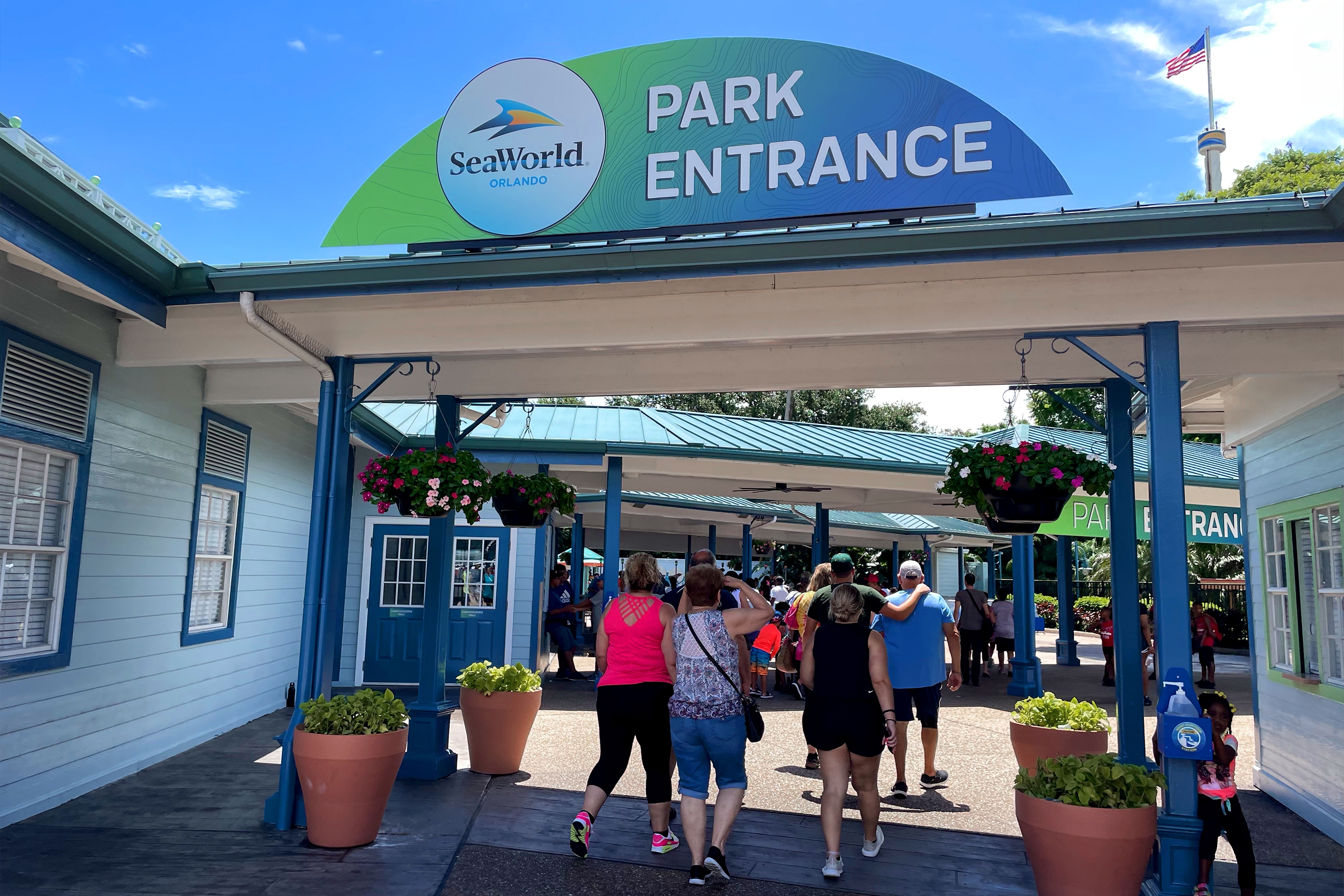 Several guests walk under the entrance queue of SeaWorld Orlando with signage that reads ‘Park Entrance’ with green and blue hues.