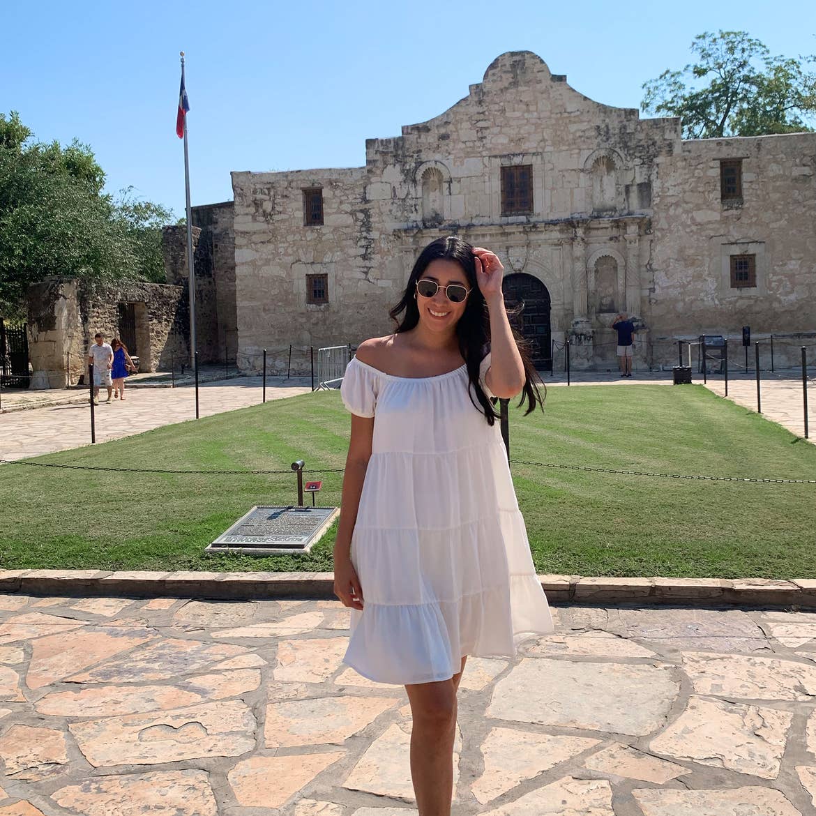 A woman in a white sundress and sunglasses stands in front of the Alamo exterior in Texas.
