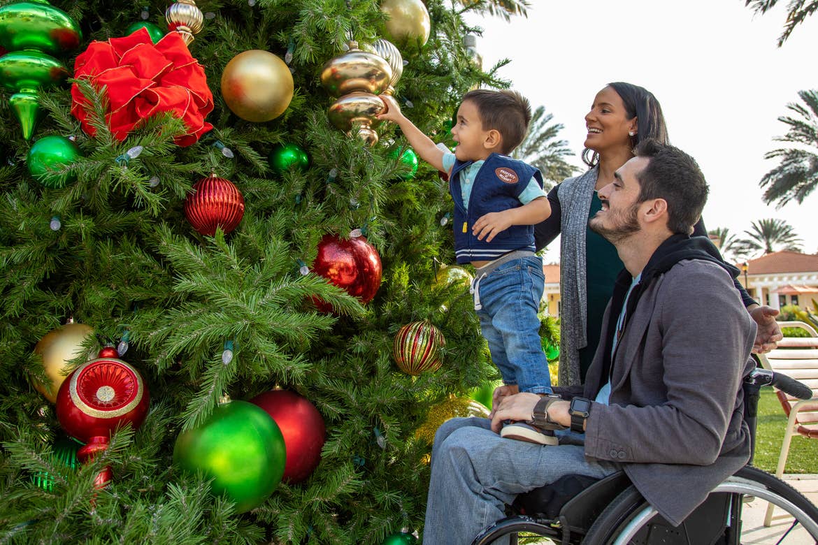 A man in a wheelchair has a little boy stand on his lap to touch large Christmas tree ornaments.