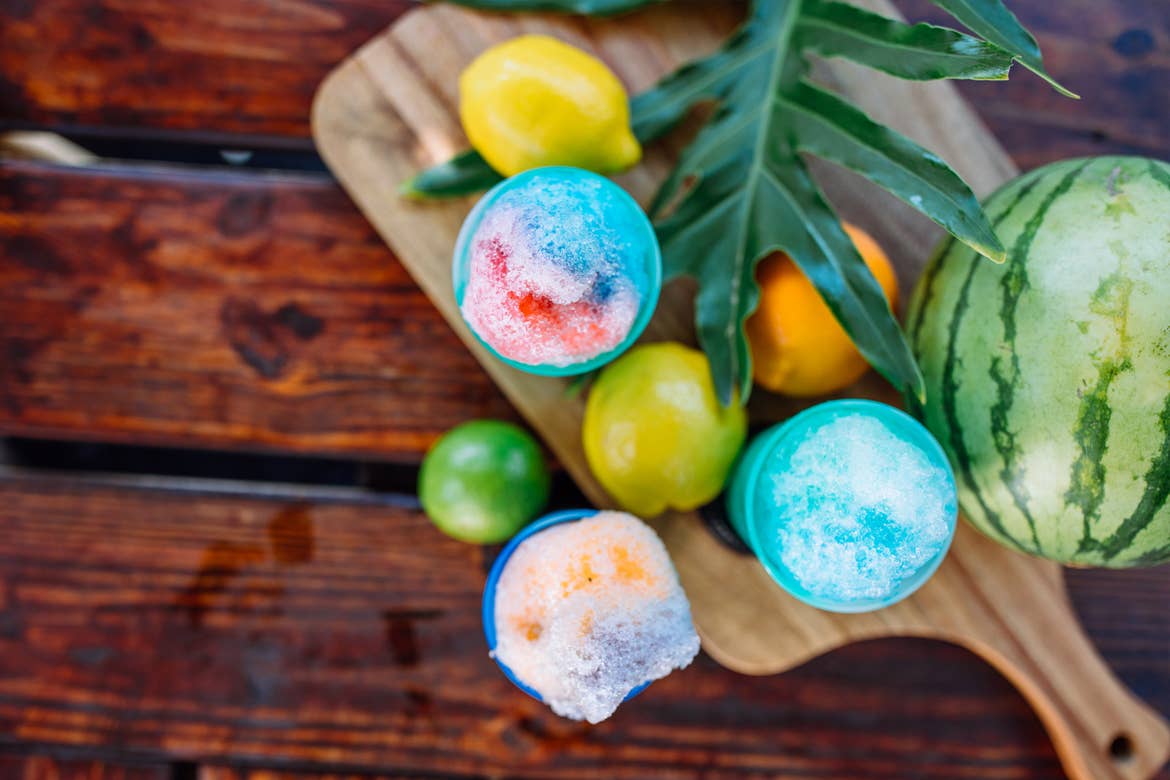 An overhead view of various colored snow cones on a wooden cutting board surrounded by citrus fruits and foliage.
