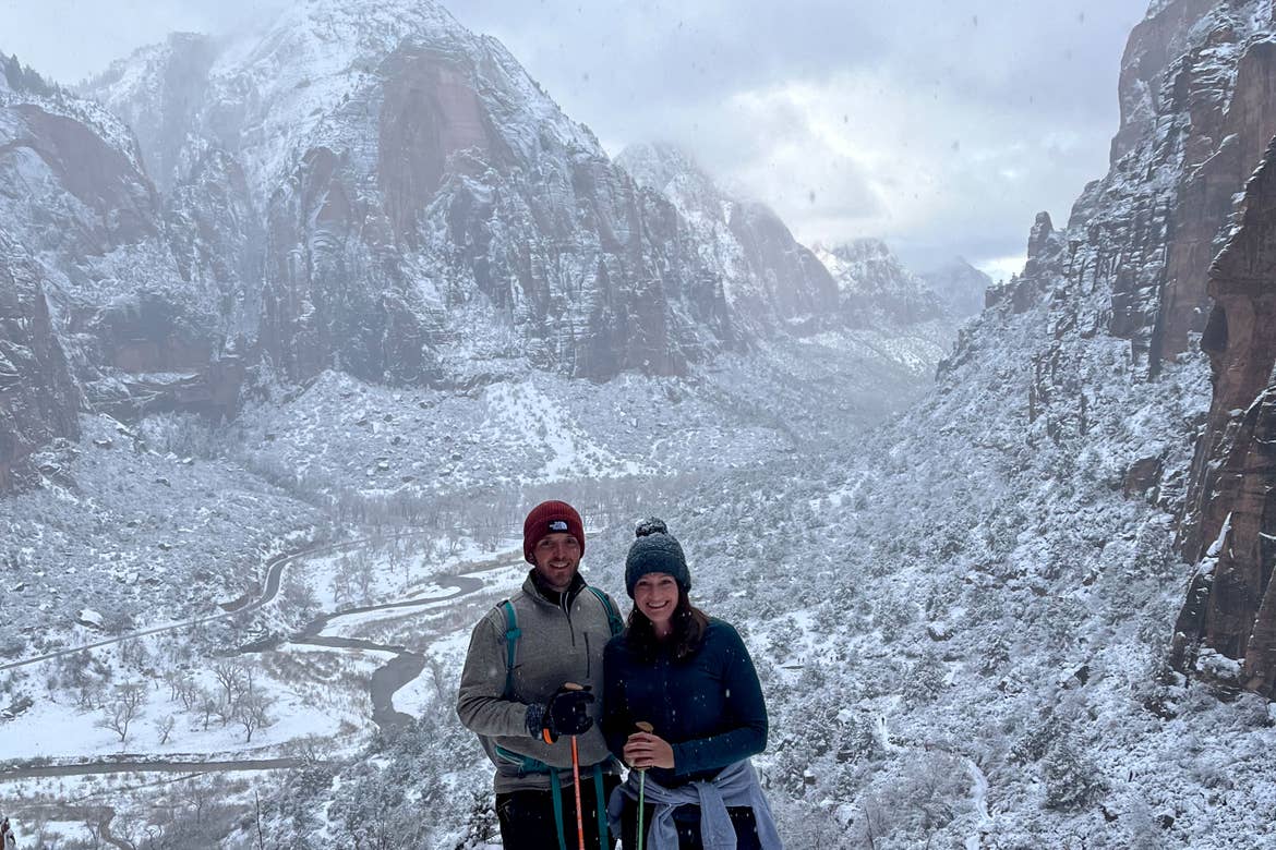 A man and woman wearing knitted hats, fleece jackets, and holding walking sticks stand in front of a snowcapped mountain range.