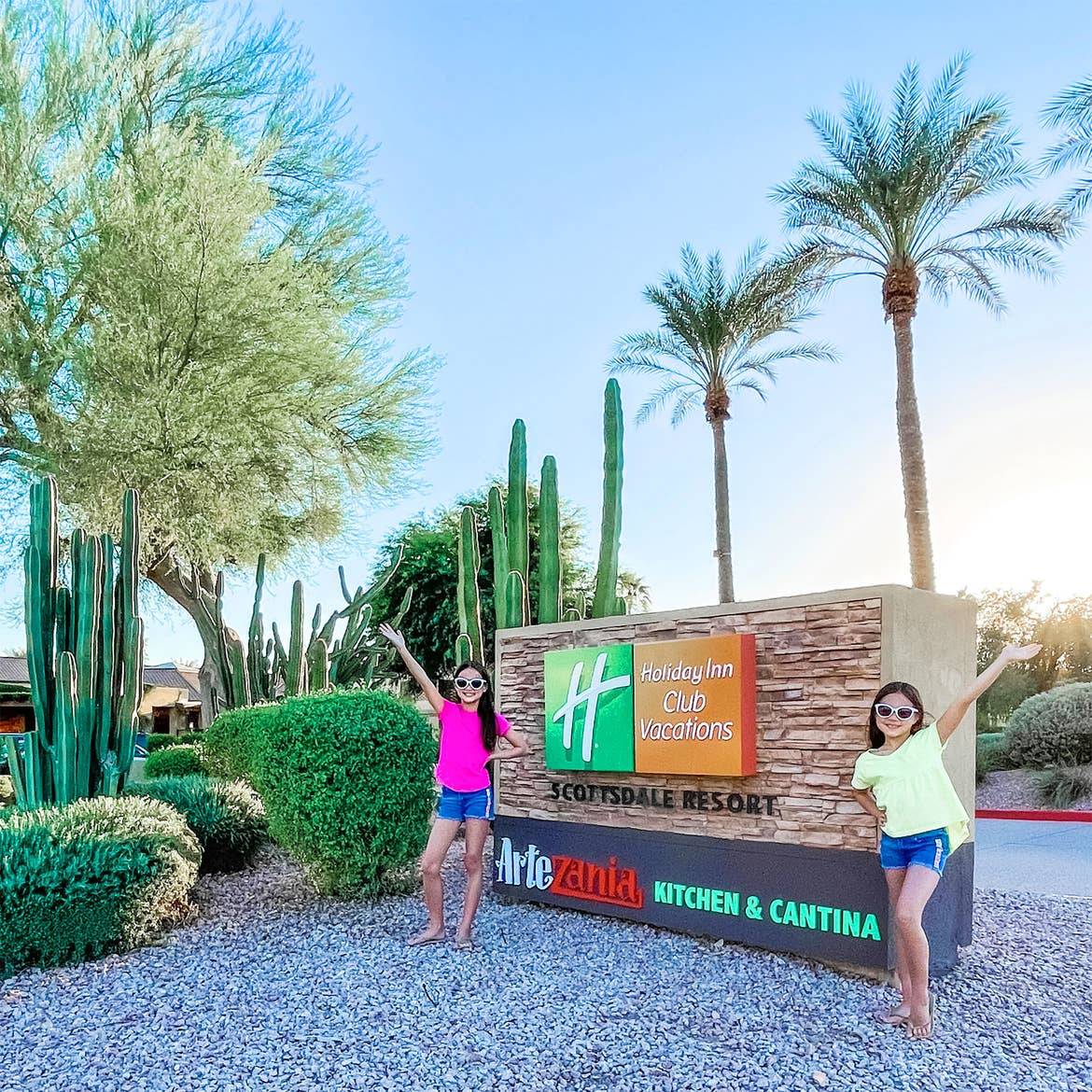 Two young girls in a pink (left) and yellow (right) t-shirt with denim shorts and sandals stand near a decorative main gate sign that reads 'Holiday Inn Club Vacations Scottsdale Resort.'