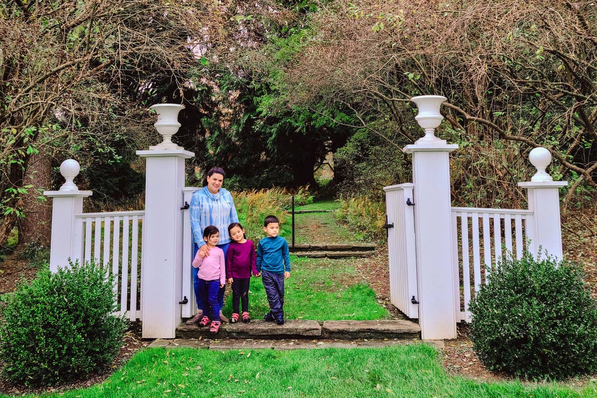 A woman and three toddlers stand in a white gateway surrounded by green landscaping.