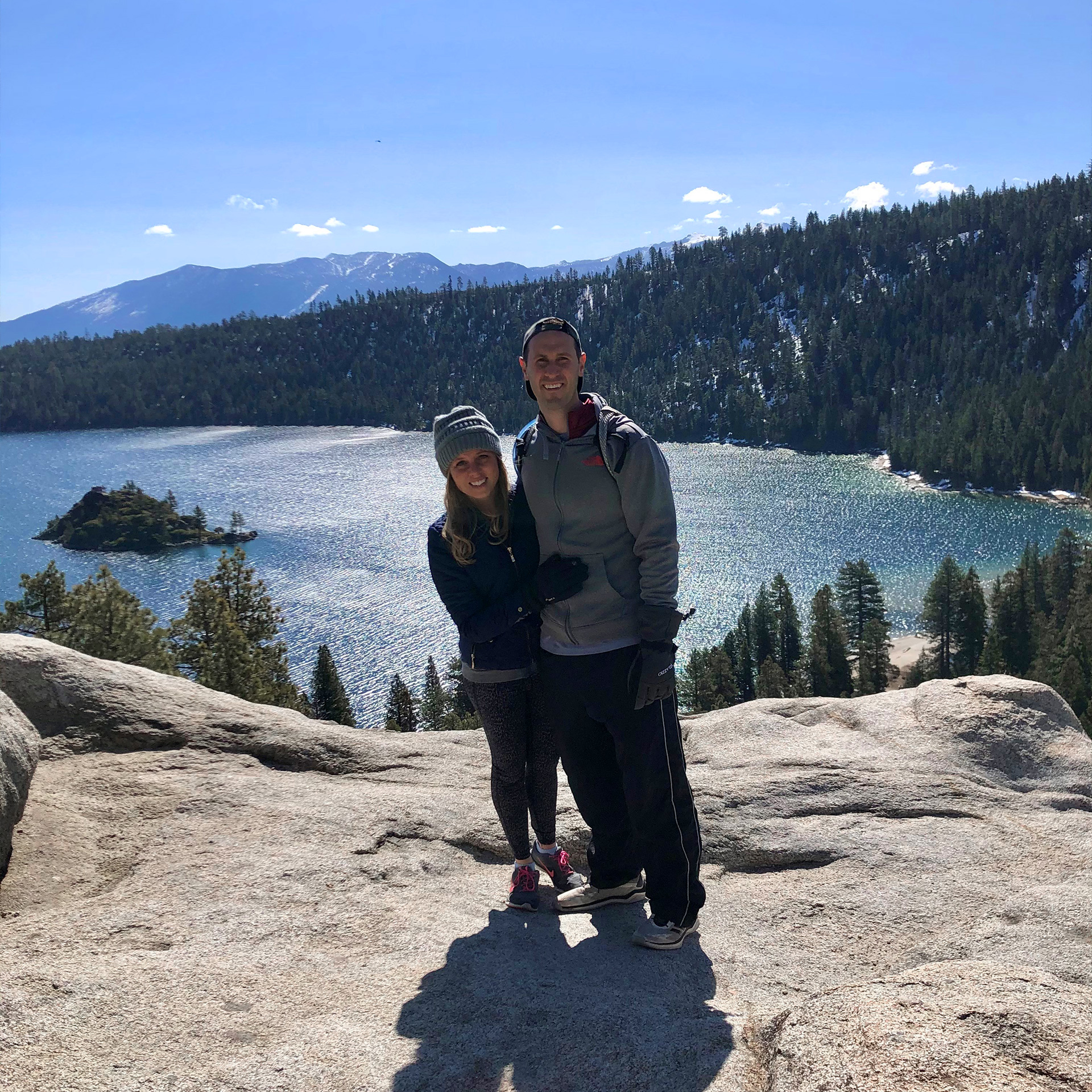 A woman (left) wears a grey beanie and black leather jacket and gloves while hugging a man (right) wearing a grey sip-up hoodie and green beanie in front of a mountain range at Lake Tahoe, Nevada.
