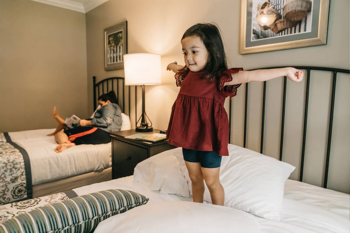 Featured Contributor, Angelica Kajiwara's daughter wears a burgundy blouse while standing and stretching on the bed in the villa of our Williamsburg resort in Williamsburg, Virginia.