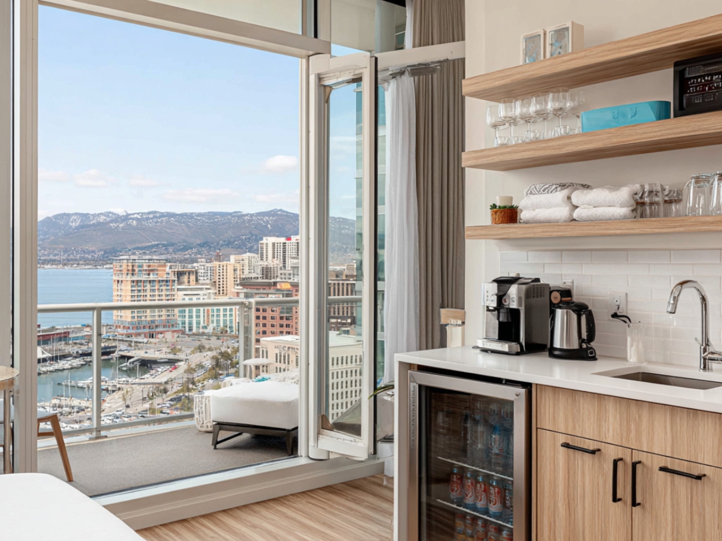 Hotel suite with floor-to-ceiling windows, kitchenette, and stunning views overlooking a city skyline and water.