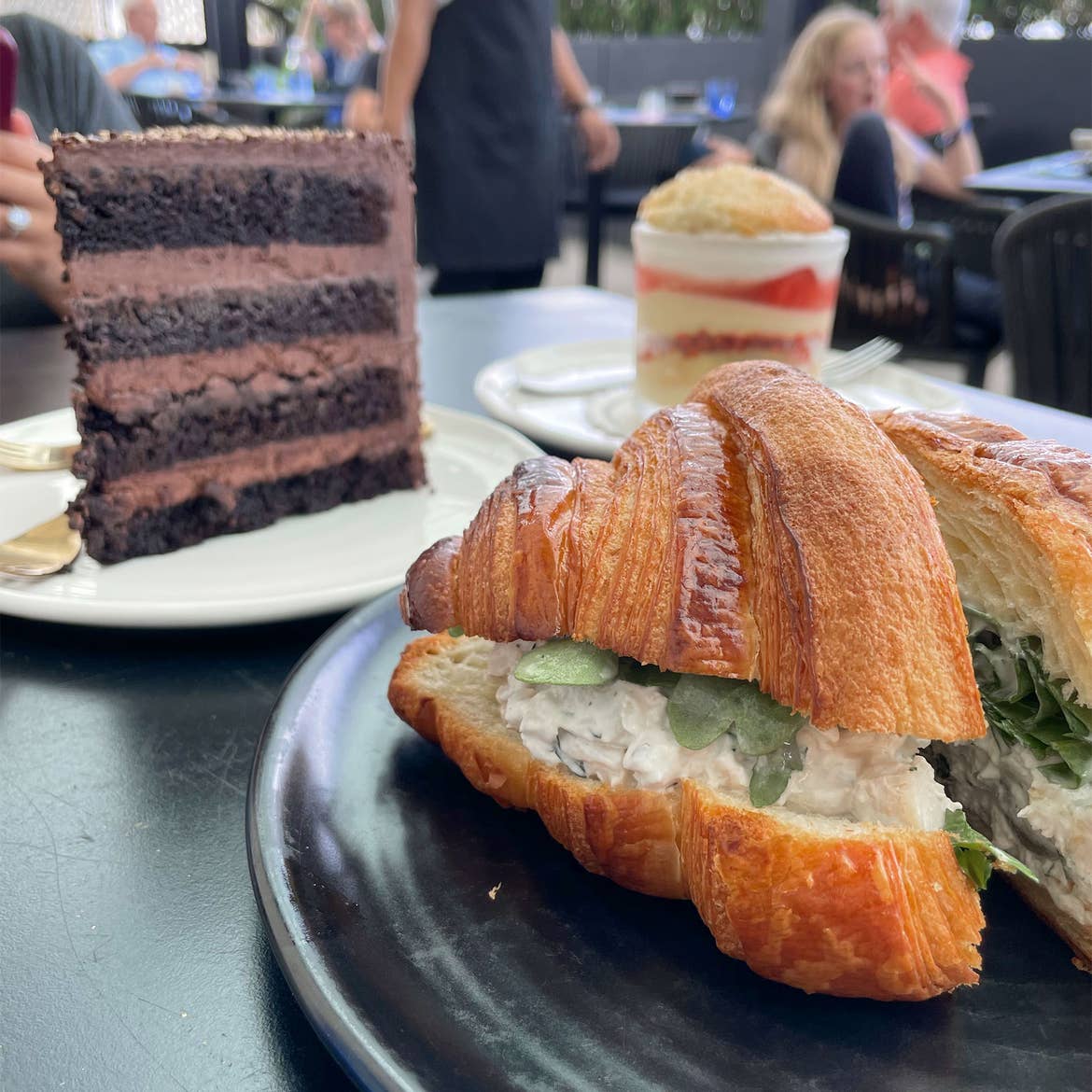 A chocolate layered cake, a parfait and a croissant sandwich are placed on various colored plates on an outdoor dining table.