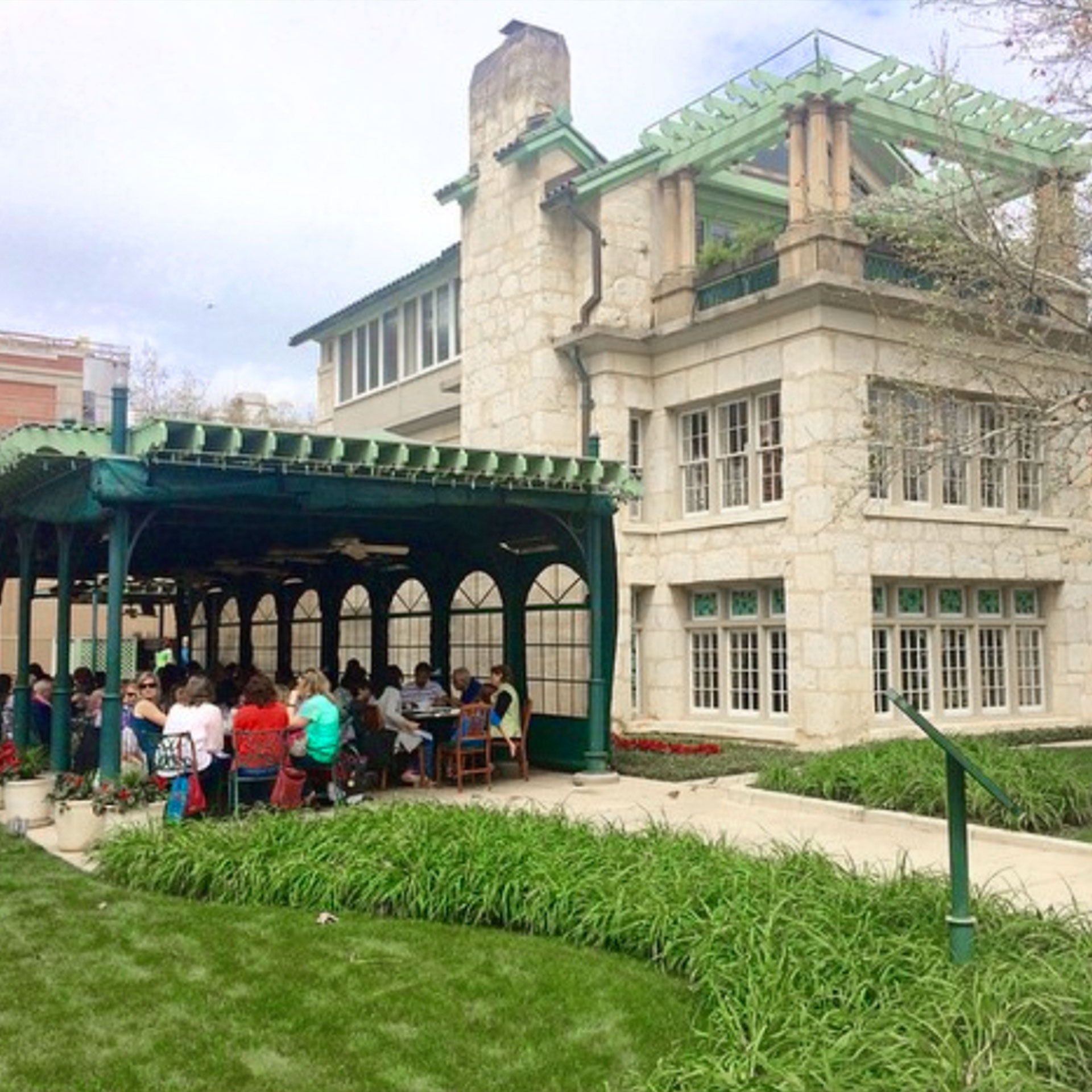 A stone-clad exterior with a green, outdoor dining patio hosts several guests near a green garden.