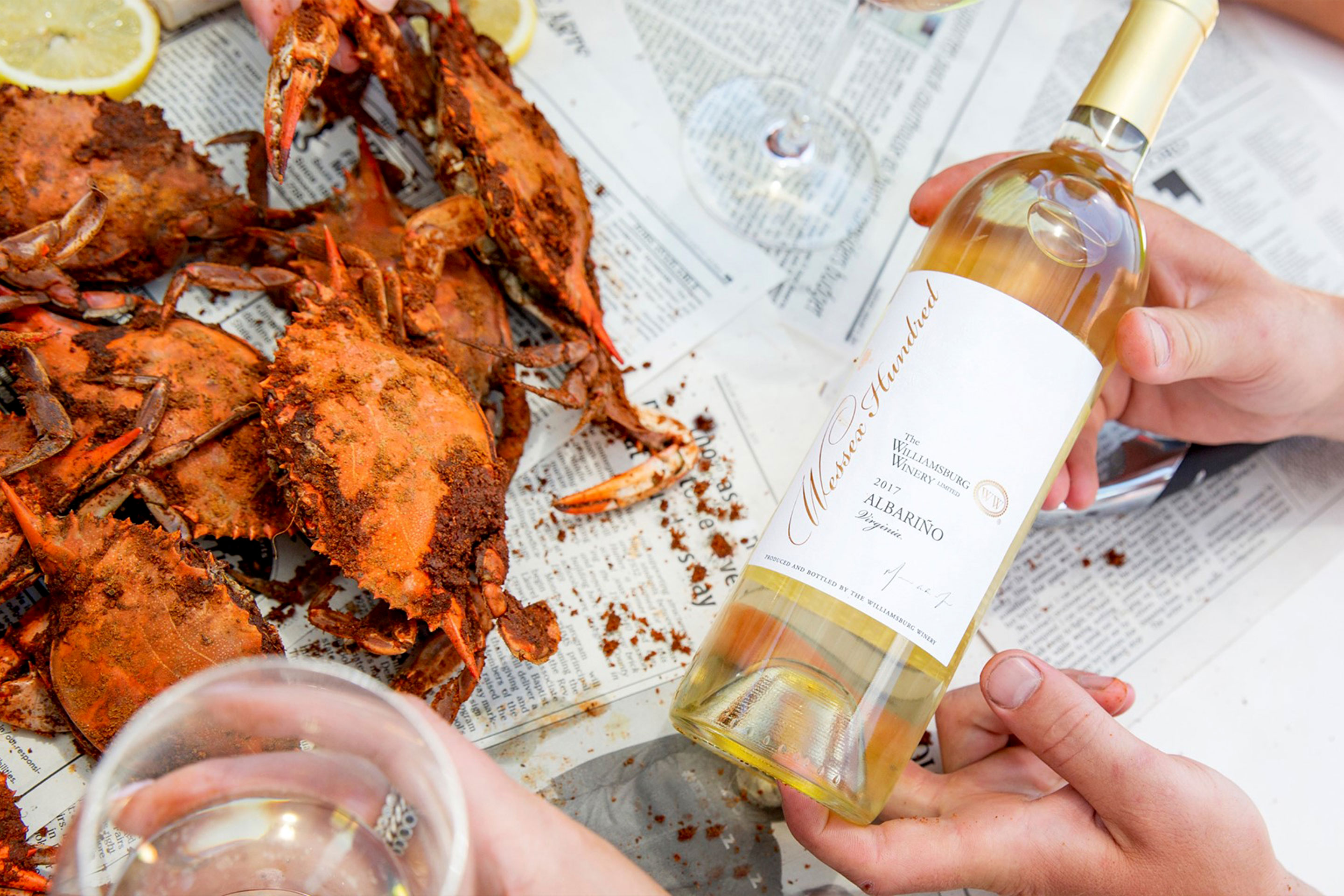A table covered with newspapers, crab, and two pairs of hands hols a glass of white wine, and its bottle.
