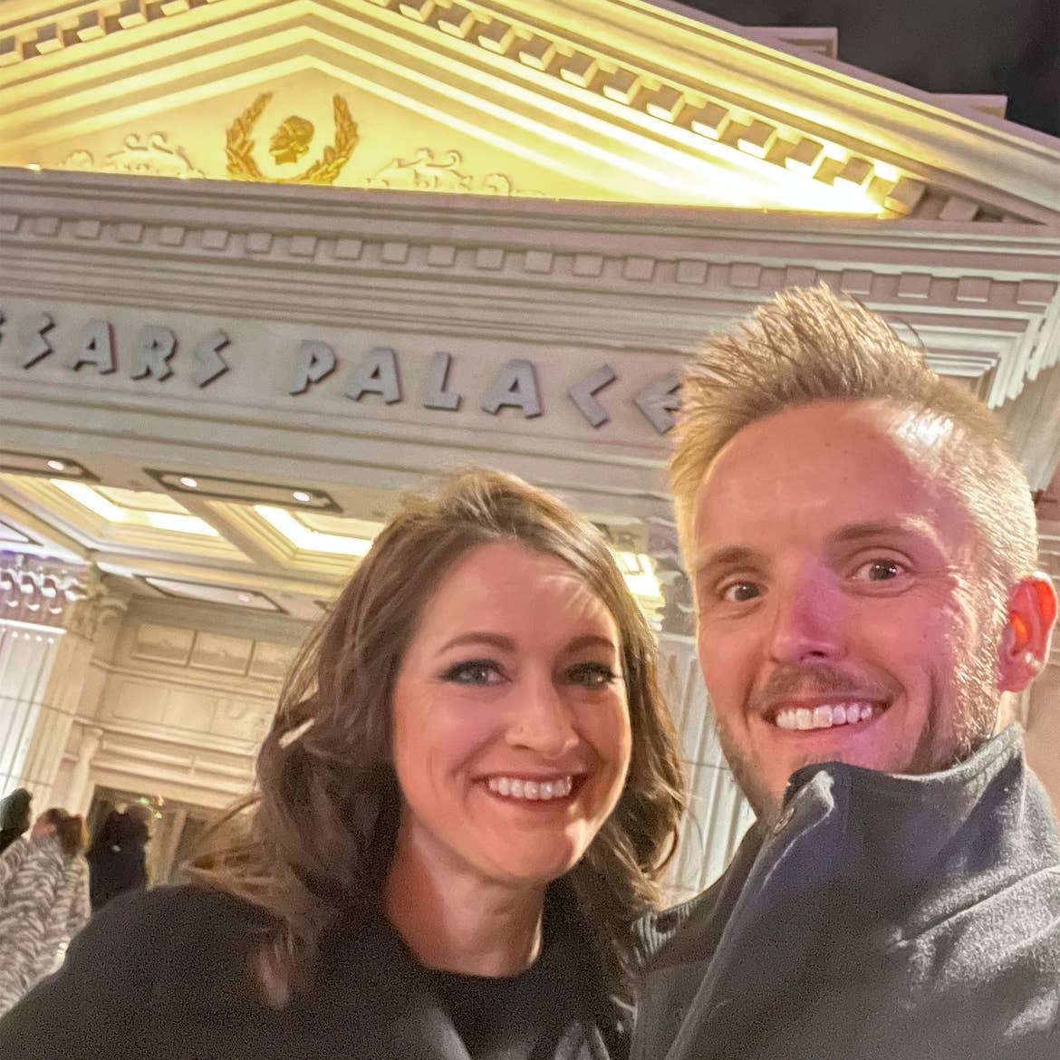 A woman and man wearing black jackets stand outside of the Caesars Palace Casino during the night.