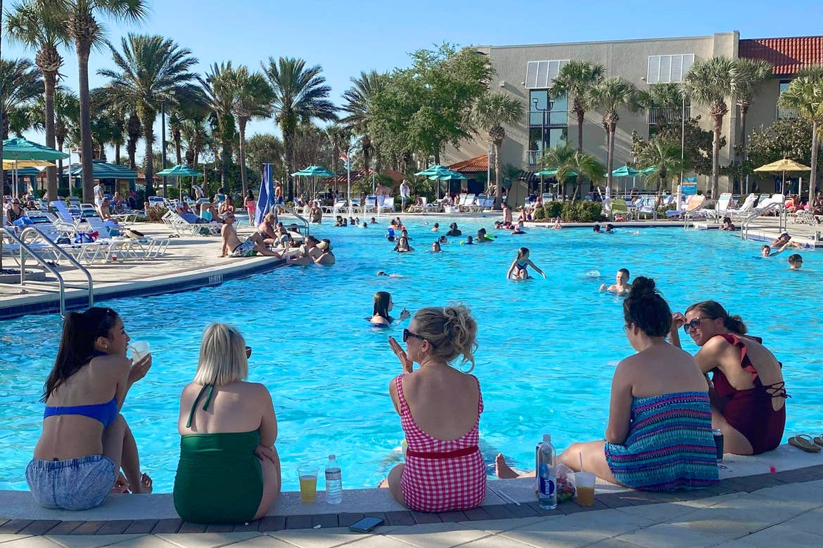 A group of caucasian women in various colored swimsuits sit at the edge of a pool surrounded by a resort facade and palm trees under a blue sky.