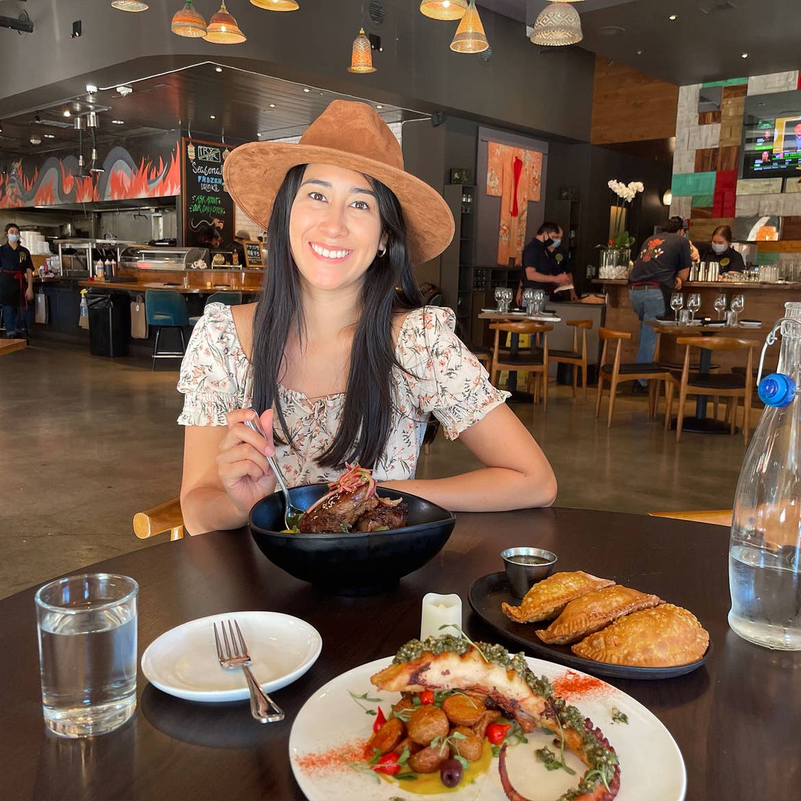 A woman in a floral sundress and sun hat sits at a wooden table with a bowl and several dishes of food in a restaurant.