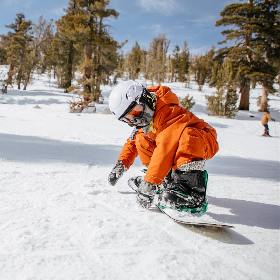 A boy wearing orange snow jacket, pants and white helmet makes his way down a snowy slopes on a snowboard.