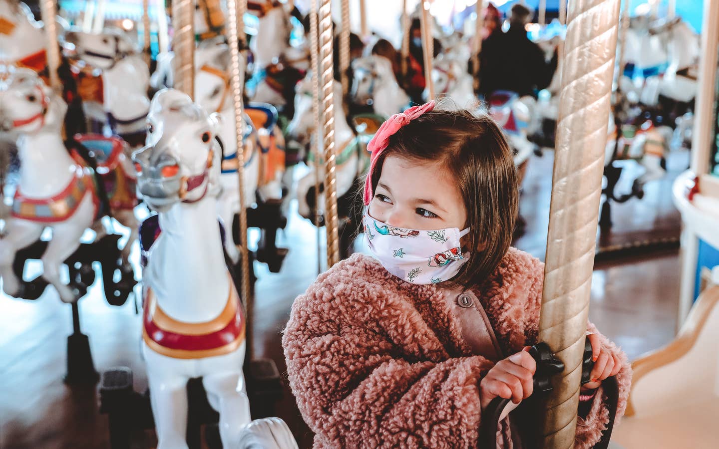 Roux rides a white horse on Prince Charming Regal Carrousel at Magic Kingdom Park at Walt Disney World® Resort wearing a pink fuzzy jacket and a 'Little Mermaid' face mask.