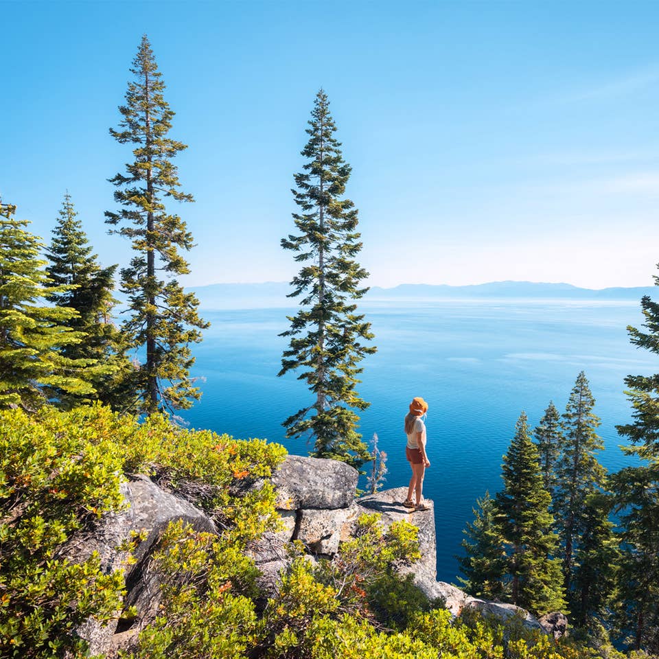 A caucasian woman wearing a white shirt, white baseball cap, shorts and boots sits on a rock at a high elevation overlooking a lake surrounded by pine trees on a mountain.