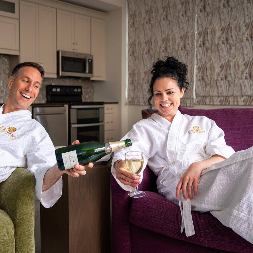 A man wearing a white robe on a green couch (left) holds a glass of champagne in his right hand while pouring the green bottle in this left hand for a woman (right) also wearing a white robe on a purple couch.