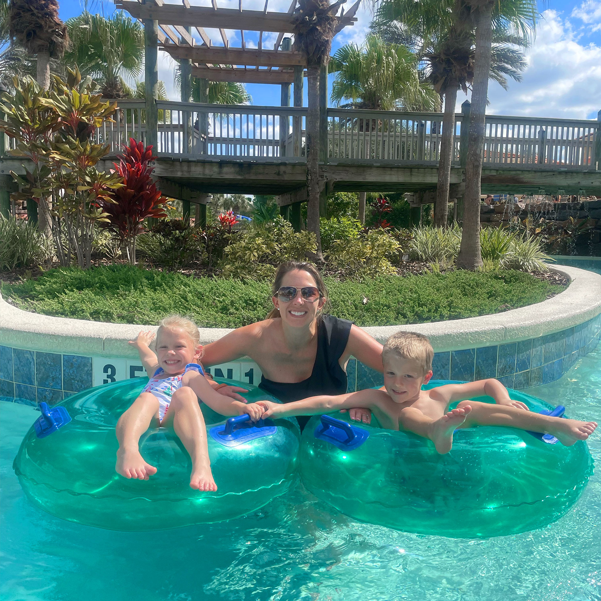 A woman in a black swimsuit and sunglasses stands in a lazy river holding a boy and girl floating in blue inflatable tubes outdoors.