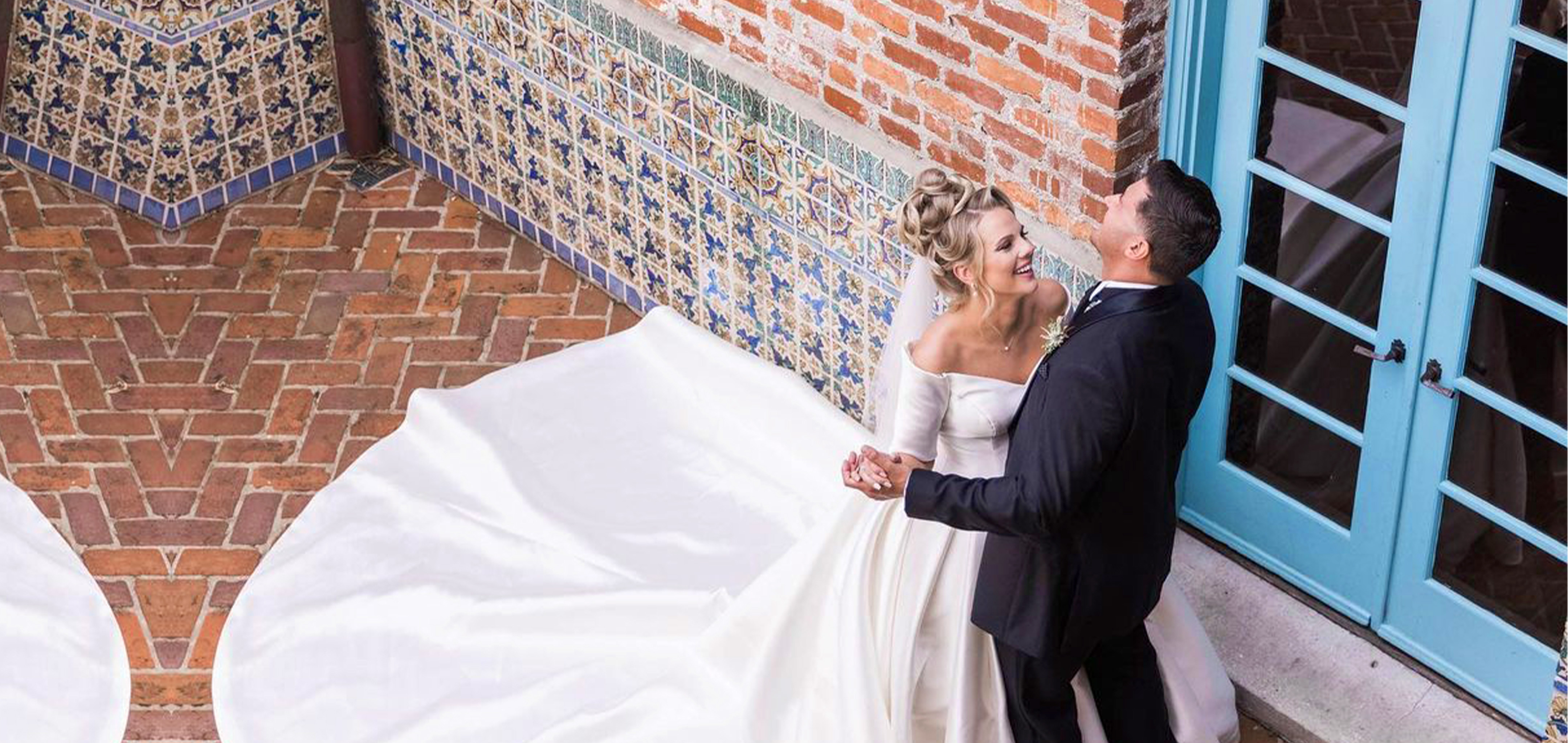 A Latinx groom wearing a black suit and (right) and a caucasian bride with a long train (left) dance with each other in front of a brick facade.