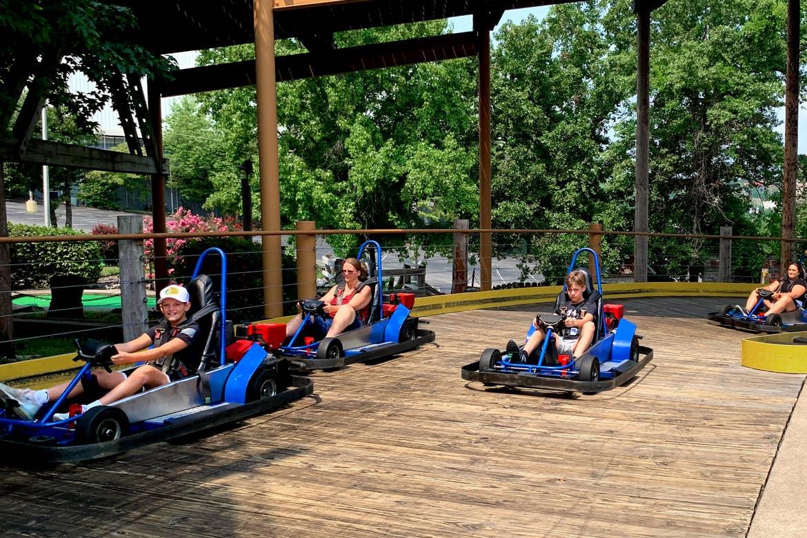 Two boys and a woman ride blue go karts on an outdoor, wooded track.