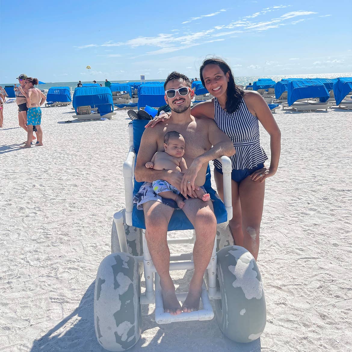 A man in a wheelchair holds an infant next to a woman on a sandy beach.