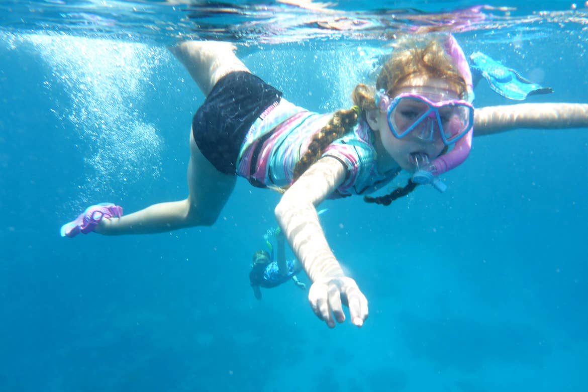 A young girl with red hair and multicolored, pastel swimwear  snorkels in the ocean.