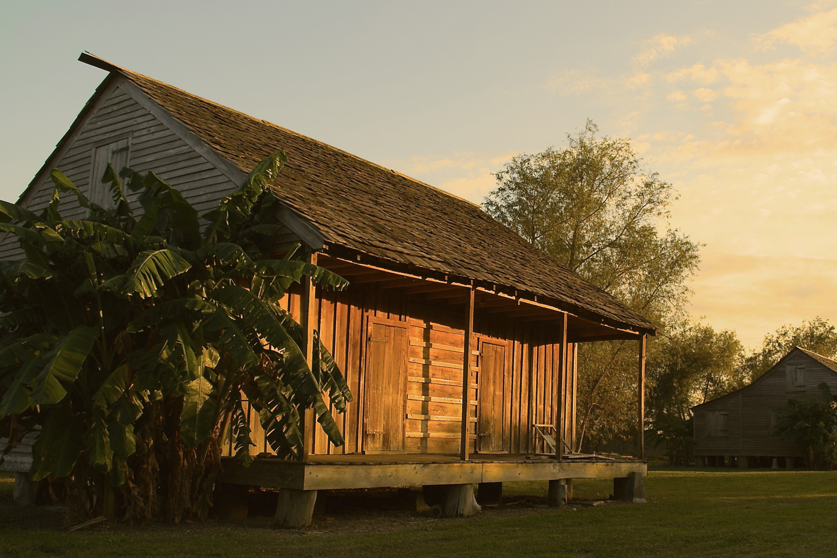 A retired wooden shack that once housed slaves at the Whitney Plantation stands vacant on a green field as the sun sets.